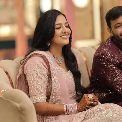 A candid shot of a happy bride at her engagement. Her makeup features a minimal, dewy finish with soft pink tones to complement her pastel pink lehenga, paired with simple wavy hair for an effortless look.