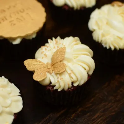 A close-up of a red velvet cupcake from the white and gold set. The design features a delicate, handcrafted golden butterfly resting on a swirl of cream cheese frosting, finished with a sprinkle of edible gold dust.
