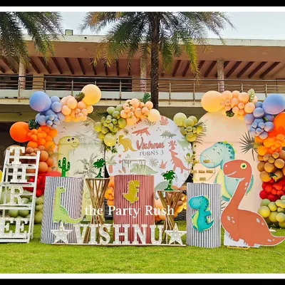 Welcome to Vishnu's Dino Park! This outdoor dinosaur-themed party is a prehistoric adventure. The setup includes multiple friendly dinosaur cutouts, a volcano-like balloon eruption, and a large 'THREE' prop, all set against a beautiful natural backdrop.