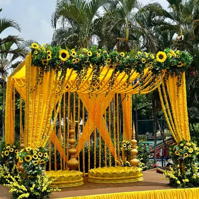 A picture-perfect Haldi mandap. The structure is draped in bright yellow fabric and adorned with strings of marigolds and sunflowers, creating a cheerful and traditional look.