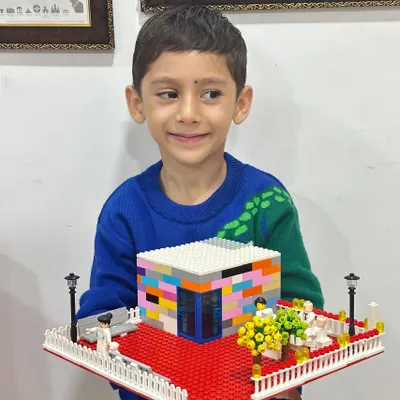 A young boy proudly holds up his impressive LEGO creation: a modern house complete with a detailed garden, fence, and a red carpet entrance. This showcases the architectural and design skills kids can develop.