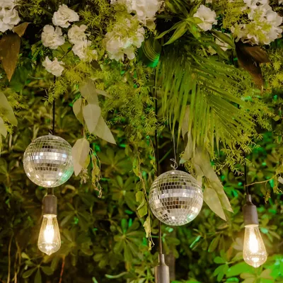 A close-up of our lighting and floral design for an evening event. We combined rustic Edison bulbs with mini disco balls and lush greenery to create a unique and magical ambiance.