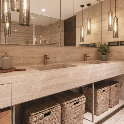 This master bathroom is a study in monochromatic luxury. A custom-built travertine vanity with integrated sinks is paired with bronze fixtures and warm pendant lighting. Wicker baskets add a natural, textural element for storage.