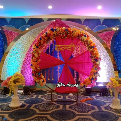 This grand Haldi stage is perfect for a banquet hall setting. The decor is centered around a large ring with 'Haldi' written in Hindi, surrounded by walls of colorful flowers.