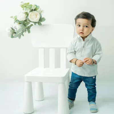 A classic pre-birthday portrait of an 11-month-old boy. Standing by a simple white chair against a white background, his serious and curious expression is the main focus of the shot.