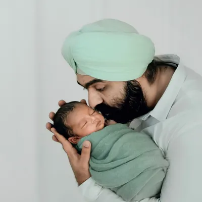 A father in a soft green turban gently kisses his swaddled newborn's head. This image celebrates the quiet, powerful bond of new fatherhood.