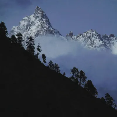 A majestic Himalayan peak emerges from the clouds at dusk. The deep blue and black tones create a moody, powerful scene that reflects the secrets the mountains hold.