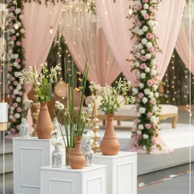 An elegant daytime event setup with a backdrop of pastel pink drapes and hanging lights. The decor is accented with pillars wrapped in pink and white roses, and terracotta vases with fresh greenery in the foreground.