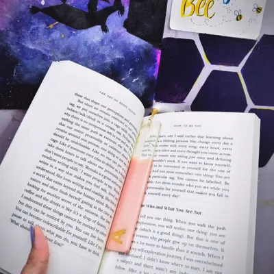A custom pastel pink bookmark with a gold 'A' initial, shown inside a book. This is how the bookmark looks in use, adding a personal touch to your reading time.