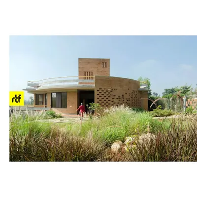 An exterior view of the 'Breathe' residence, showcasing the curved Jaali wall made of compressed earth blocks. The surrounding native grasses and plants are not just decoration but an integral part of the home's ecosystem.