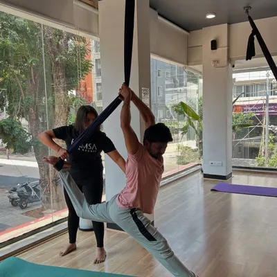 An instructor provides a hands-on assist to a student in a supported split pose during an aerial yoga class.