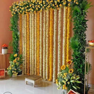 Here is a classic Haldi function backdrop I created, using vertical strings of yellow and white marigolds. The frame is bordered with fresh green foliage, and I've placed a traditional wooden chowki in front for the ceremony. This design is perfect for creating a vibrant, traditional atmosphere at home.
