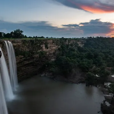 The elements of nature combine in this long-exposure shot of a waterfall at sunset. The image captures the motion of the water, the colors of the sky, and the lushness of the surrounding forest.