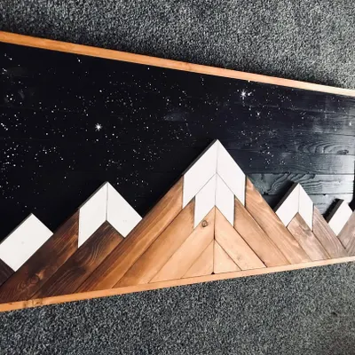 This rustic wooden wall art captures a mountain range against a starry night sky. The contrast between the natural wood grain of the mountains and the dark, speckled sky is stunning.