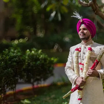 A classic portrait of the groom in his sherwani and magenta turban, holding the ceremonial sword. He looks regal and ready for his wedding.