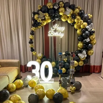 A sophisticated 30th birthday setup with a black and gold circular balloon arch, a 'Happy Birthday' neon sign, and a '30' marquee light. A photo display stand adds a personal touch.