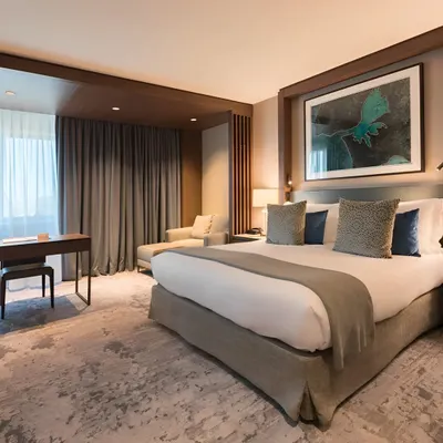 This luxury hotel suite design balances comfort with sophistication. The room features a plush king-sized bed, a dedicated work desk, a comfortable chaise lounge by the window, and curated artwork, all tied together with a calming, neutral color palette.