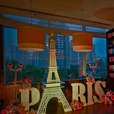 Bringing the romance of Paris to a party with this stunning setup. It features a large Eiffel Tower cutout, 'PARIS' marquee letters, and beautiful floral arrangements, all set against a city skyline view.