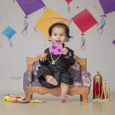 A happy baby in a black kurta set for Makar Sankranti, holding a small kite against a backdrop of colorful patangs. This is a fun and festive baby photoshoot.