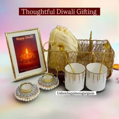 The 'Golden Glow' Diwali basket, featuring two elegant white and gold mugs, matching spoons, decorative pearl diyas, and a 'Happy Diwali' photo frame. This hamper is perfect for couples.