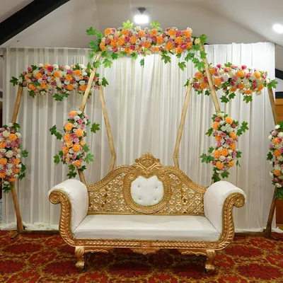 This premium rental includes an elegant gold and white two-seater sofa, set against a white fabric backdrop and framed by a beautiful floral arch structure. It is ideal for an engagement or reception stage.