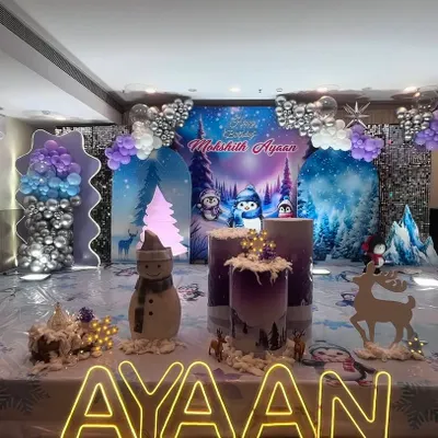 This is a full view of a 'Winter Wonderland' themed birthday stage I designed. The decor features a snowy forest backdrop, balloon columns in purple, white, and silver, and custom props including a light-up name sign for 'Ayaan'.
