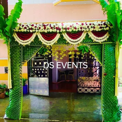 A classic South Indian wedding entrance. This arch is constructed with a frame of woven coconut leaves, decorated with red and white floral thoranams, and flanked by auspicious banana plants.