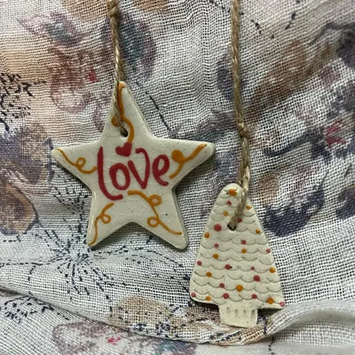 A set of 'All About Love' ornaments, featuring a star and a Christmas tree. The red and orange details are hand-painted, making each piece one-of-a-kind.