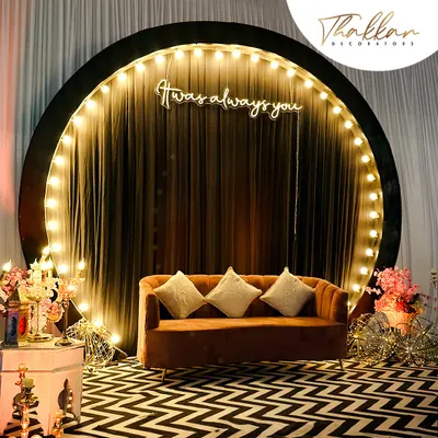 A modern and chic photo booth for a reception. The setup features a circular backdrop with a "It was always you" neon sign, a stylish sofa, and a trendy zig zag floor.