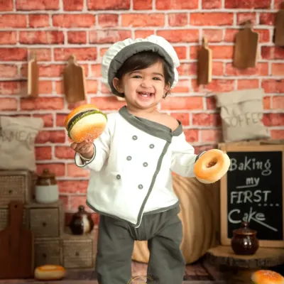 The 'Little Chef' is ready to cook! This happy toddler is holding a toy burger and donut in our custom kitchen set, complete with a brick wall and baking props.