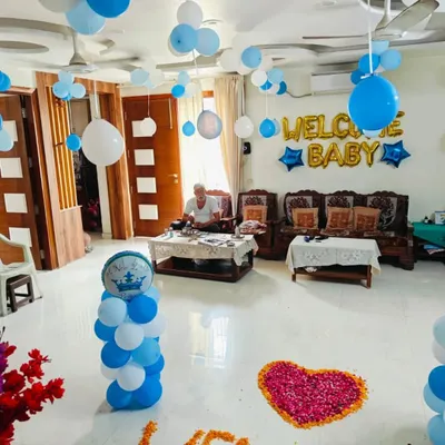 A heartwarming "Welcome Baby" decoration for a home. The setup includes foil letter balloons, balloon bunches hanging from the ceiling, and a flower petal heart on the floor.