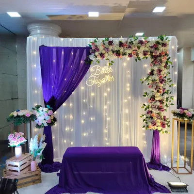 An elegant engagement backdrop with a "Better Together" neon sign, featuring purple and white drapes, a floral arch, and fairy lights.