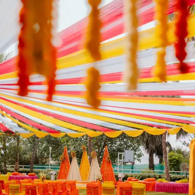 A full view of an outdoor Haldi setup, showcasing the vibrant canopy of red, yellow, and white fabric strips. The seating area and floral backdrops are all coordinated to create a bright and joyful environment for the pre-wedding festivities.