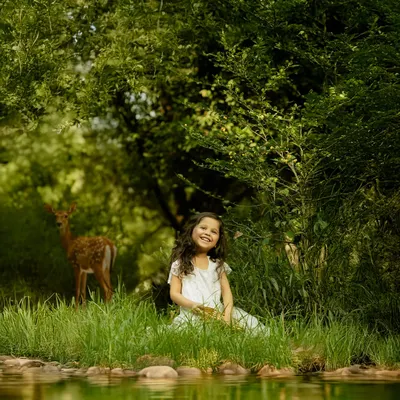 A young girl in a white dress sits by a serene pond, with a deer visible in the lush green background. This magical, storybook-like image showcases the beauty and unpredictability of outdoor photography.