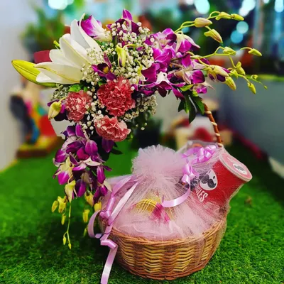 This stunning gift basket combines a bouquet of fresh flowers with a selection of goodies. The arrangement includes lilies and orchids alongside a neatly wrapped package of treats, creating an elegant and versatile gift for any special day.