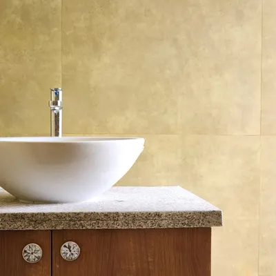 A close-up of a bathroom vanity, featuring a simple white vessel sink on a granite countertop. The brass-toned wall tiles add a warm, metallic sheen, complemented by unique, decorative cabinet knobs.