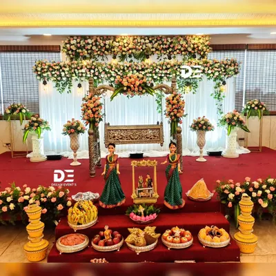 An elegant naming ceremony setup featuring a beautifully carved wooden cradle, abundant fresh flowers, and traditional brass lamps. The display of fruits and sweets adds to the richness of the occasion.