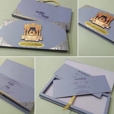 This premium boxed invitation in a sophisticated grey-blue features a 3D metallic emblem of a wedding ceremony. The set includes a matching carry bag and multiple inserts for a complete luxury experience.