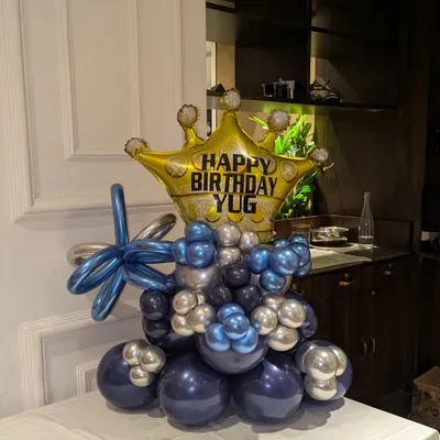 A royal-themed balloon bouquet featuring a large gold crown foil balloon, personalized with the name 'YUG'. The base is made of blue and silver chrome balloons.