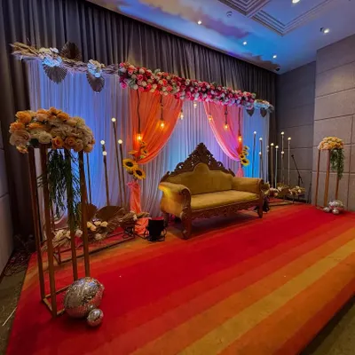 An elegant stage for an anniversary celebration. The decor features a floral runner of pink and white roses, peach-colored drapes, hanging Edison bulbs, and a classic carved sofa.
