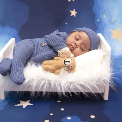 A perfect picture of peaceful sleep. This newborn is dressed in a cozy blue sleeper, cuddling a teddy bear while napping on a miniature white bed against a starry night backdrop.