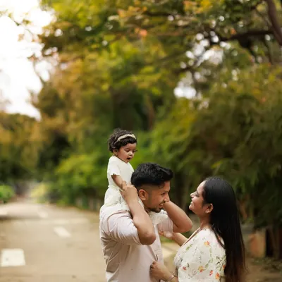 The partnership and joy in parenting shine through in this outdoor family portrait. A father carries his daughter on his shoulders while a mother looks on with love.