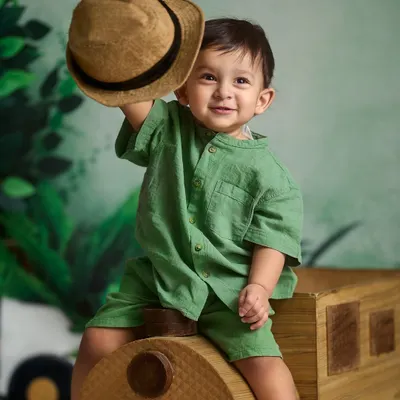 A charming tip of the hat from this little gentleman. This indoor portrait was taken against a jungle-themed backdrop with a wooden train prop, showcasing a fun and creative studio setup.