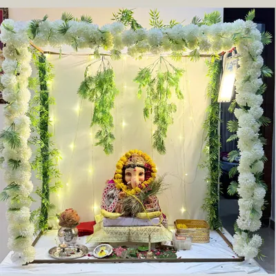 A serene and elegant Ganesh Chaturthi decor for your home. This setup features a simple frame draped with white floral garlands and hanging greenery, illuminated by fairy lights for a divine glow.