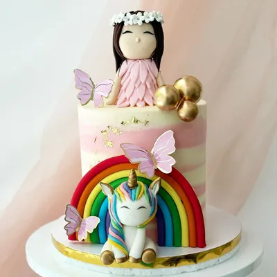 A magical unicorn and fairy theme cake. This design features a rainbow, a cute unicorn, a fairy girl topper, and delicate butterfly accents on a pink striped buttercream base.