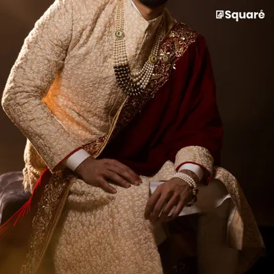 We craft elegant sherwanis that are custom-fitted to make you look and feel exceptional on your special day. This textured beige sherwani, paired with a deep red velvet shawl and pearl accessories, exemplifies the regal aesthetic we deliver for discerning grooms.