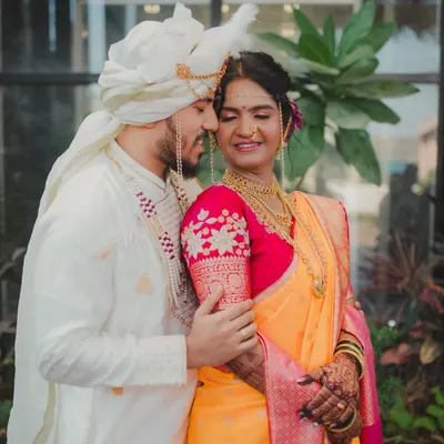 A classic portrait of a Maharashtrian couple, capturing the groom's pride and the bride's gentle smile. The groom is dressed in a traditional white kurta and Pheta, complementing the bride's vibrant yellow and pink Paithani saree.
