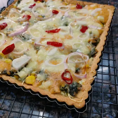 A freshly baked rectangular Corn and Spinach Quiche, with a golden-brown crust, cheesy filling, and topped with rings of onion and red chili.