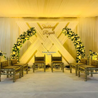 This engagement stage features a unique backdrop of crisscrossing white drapes and warm gold accents. The design is completed with two beautiful arches of yellow flowers and traditional bajot-style seating for the couple.