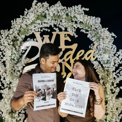 A fun and creative save-the-date photo where a couple holds up custom newspapers announcing their wedding date in front of a floral arch.